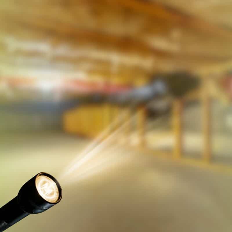 Flashlight shining a beam of light into a dimly lit crawl space with wooden support framing.