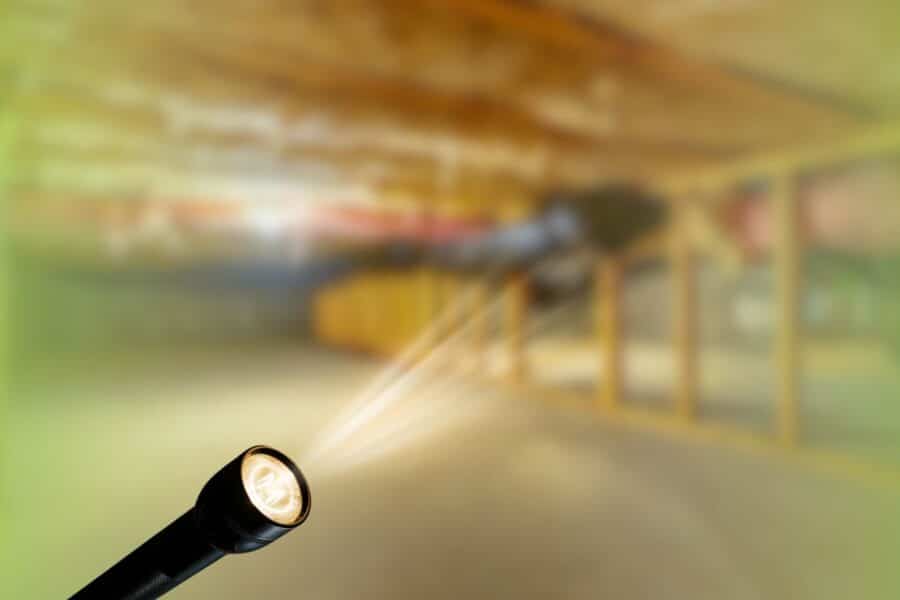 Flashlight shining a beam of light into a dimly lit crawl space with wooden support framing.
