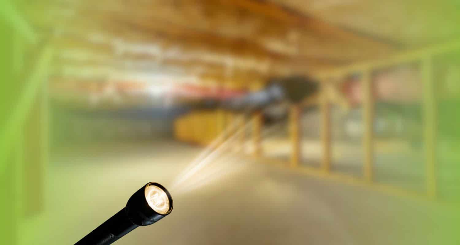 Flashlight shining a beam of light into a dimly lit crawl space with wooden support framing.